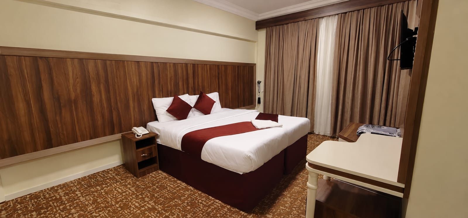 A hotel room with a wooden headboard, a bed with white bedding and red accents, a bedside table with a phone, a small nightstand, a desk with a chair, and a large window with curtains.