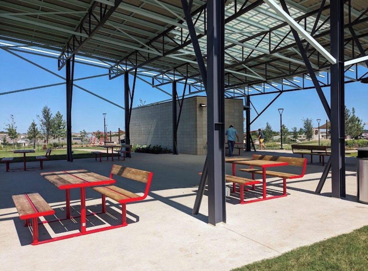 An outdoor covered seating area with benches and tables, a trash can, and a building in the background.