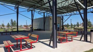 An outdoor covered seating area with benches and tables, a trash can, and a building in the background.