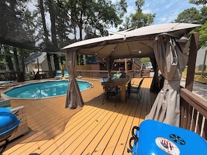 Una terraza de madera con piscina, un área de estar cubierta y sillas de descanso.