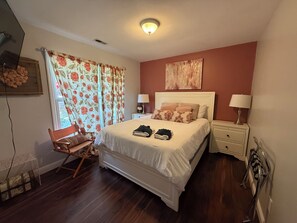 Un dormitorio con una cama, dos mesitas de noche, una silla, una ventana con cortinas y una lámpara en el techo.