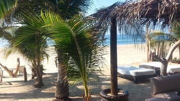 Un espace de détente aménagé au bord de la plage, avec des palmiers, un toit de chaume et des sièges en plein air.