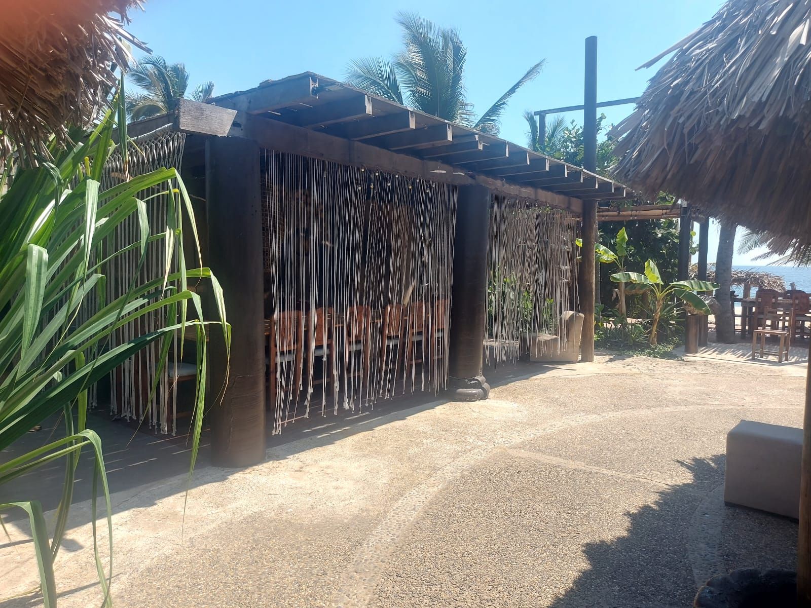 Un espace extérieur aménagé avec des sièges, abrité par un toit de chaume, entouré de plantes tropicales et recouvert de gravier.