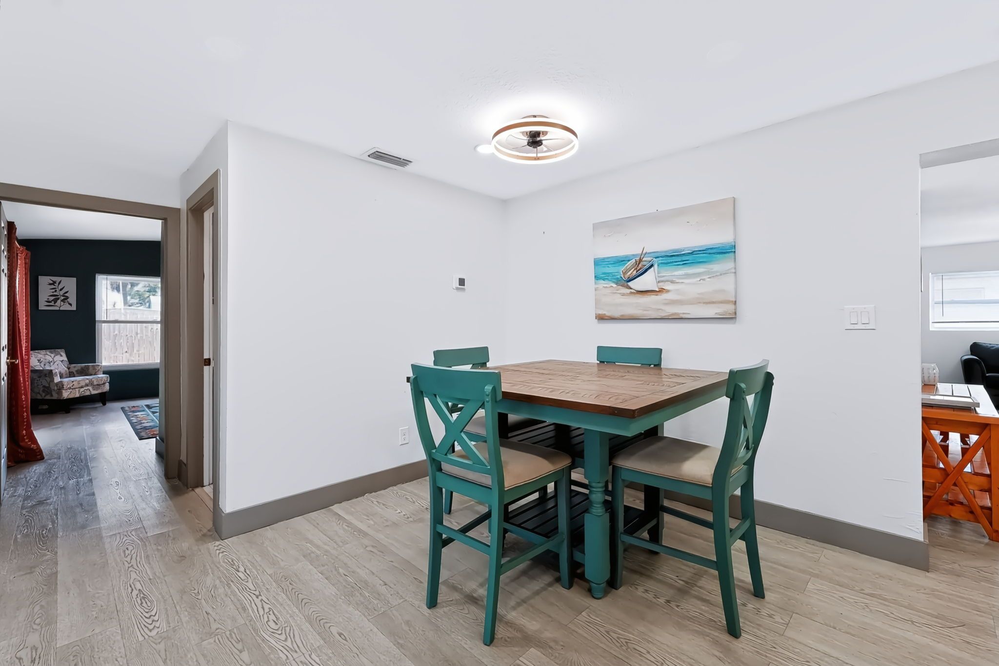 A dining area with a wooden table and teal chairs, a ceiling fan, and a beach-themed painting on the wall.