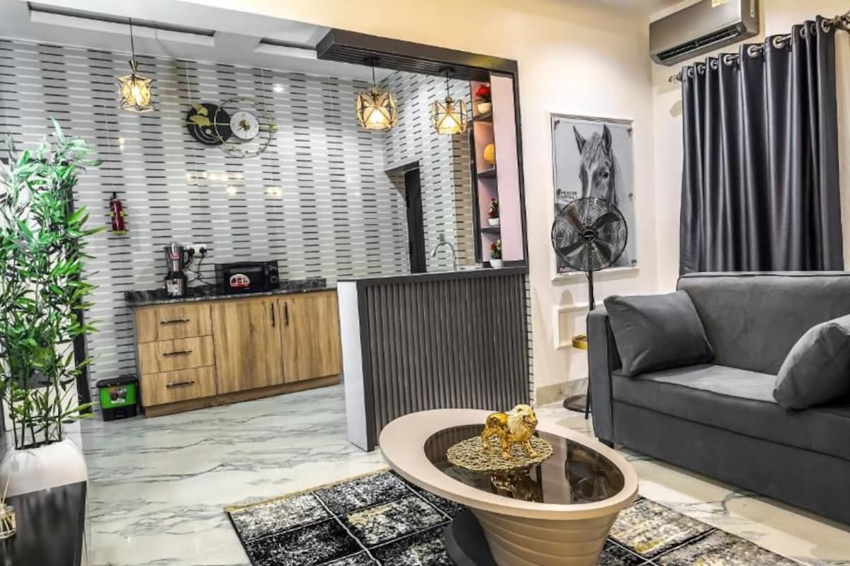 A modern living room with a grey sofa, a coffee table with a decorative item, a kitchen area with wooden cabinets, and a wall with a circular mirror and artwork.