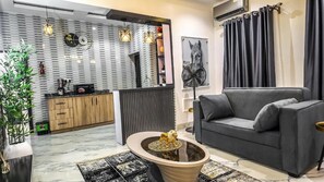 A modern living room with a grey sofa, a coffee table with a decorative item, a kitchen area with wooden cabinets, and a wall with a circular mirror and artwork. - Jayhaven Crystal Serviced Apartment (Uyo)