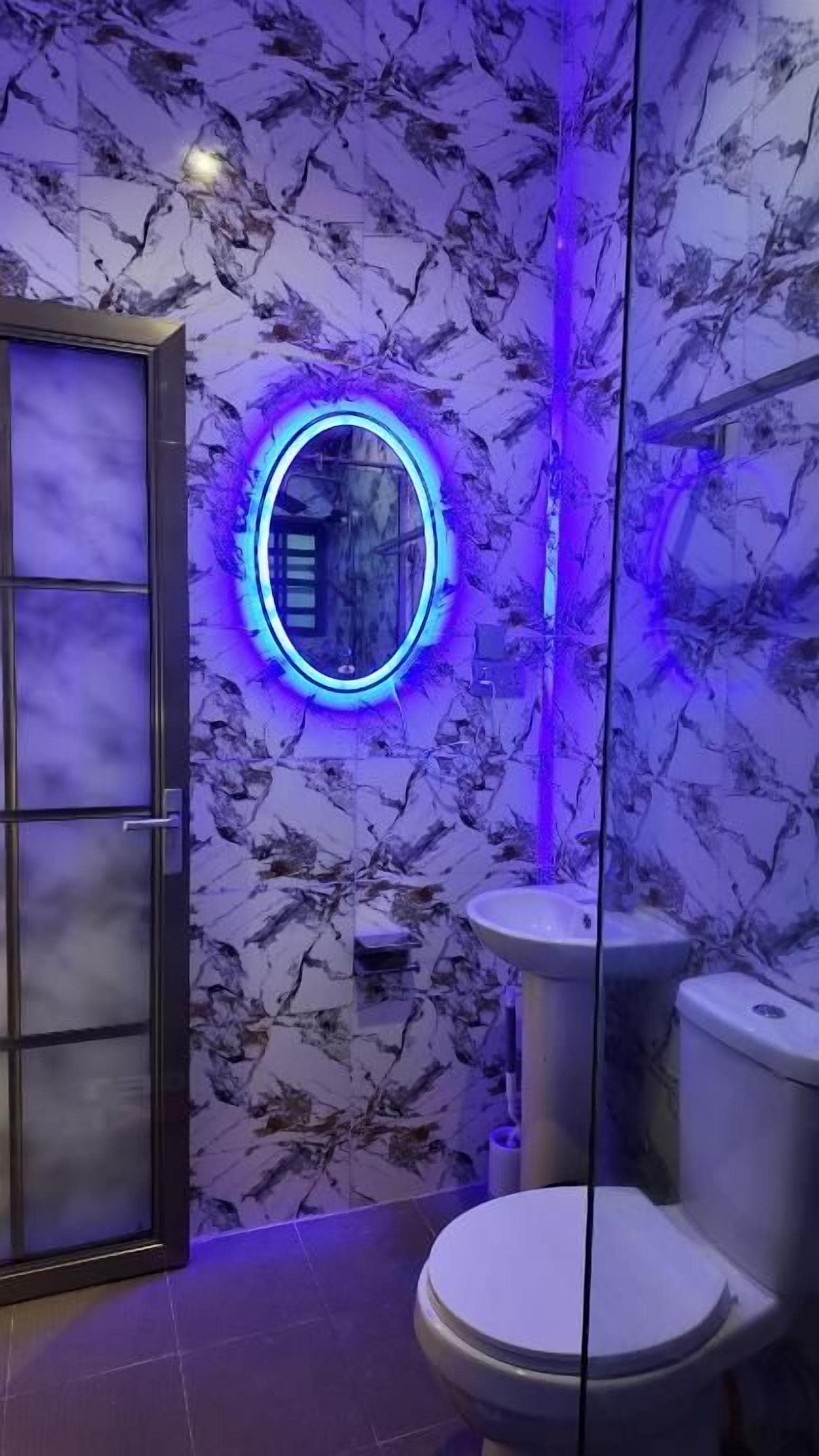 Ein Badezimmer mit Marmorwand, einem runden Spiegel mit blauer Beleuchtung, einer Toilette und einer Duschkabine aus Glas.