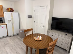 Cozinha compacta com mesa de jantar, cadeiras, geladeira, lixeira, armário com televisão e uma porta.