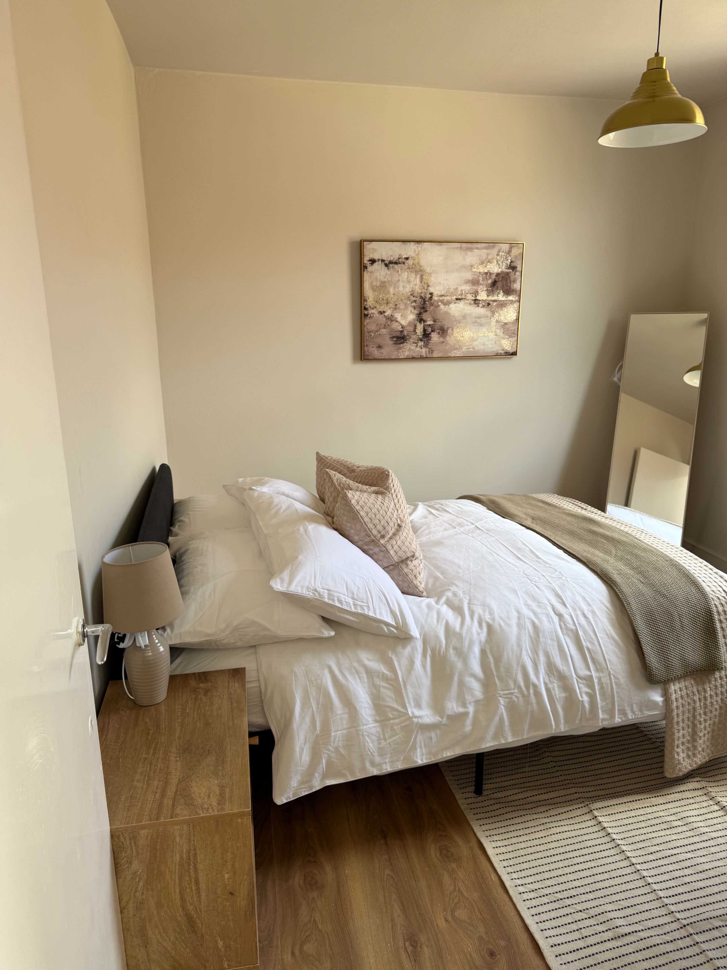 Un dormitorio con cama, almohadas, una lámpara de noche, un cuadro abstracto en la pared y un espejo.