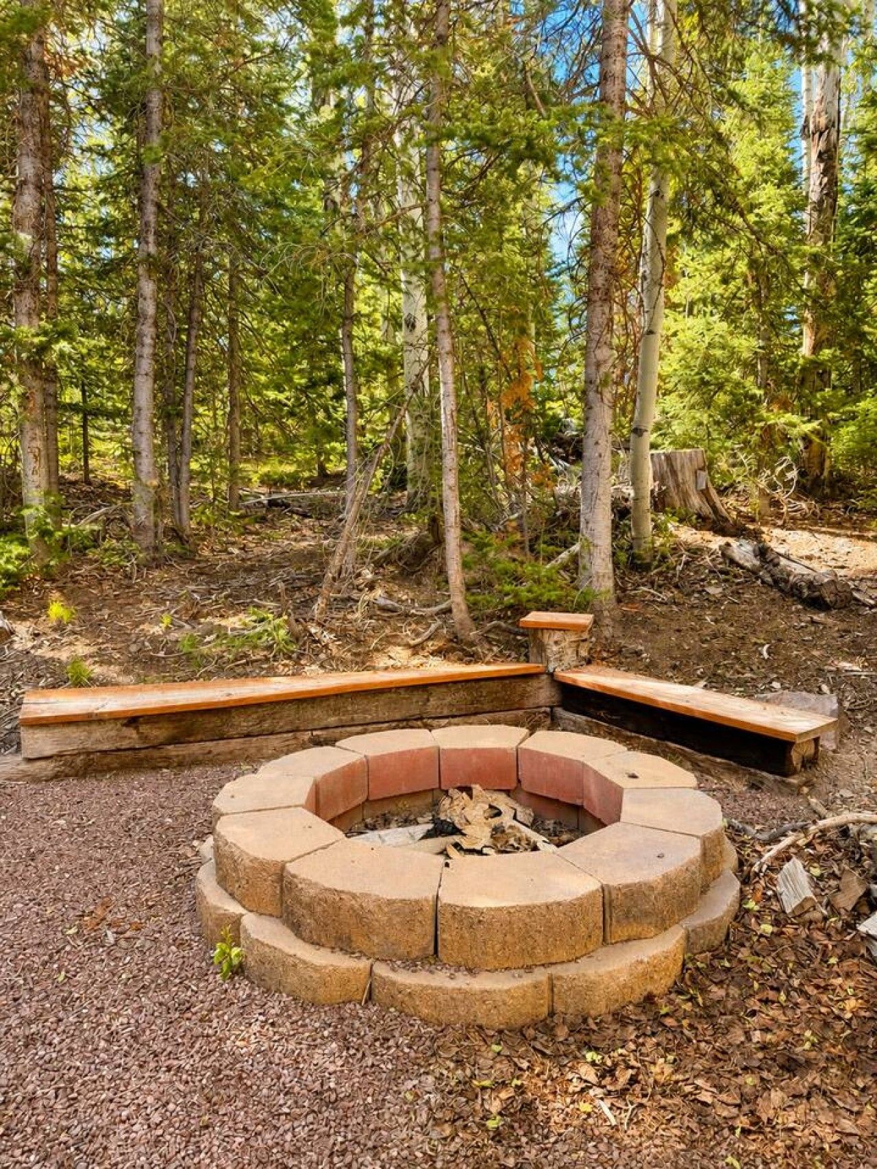 A circular fire pit surrounded by stone seating, set in a forested area with trees and a clear sky.