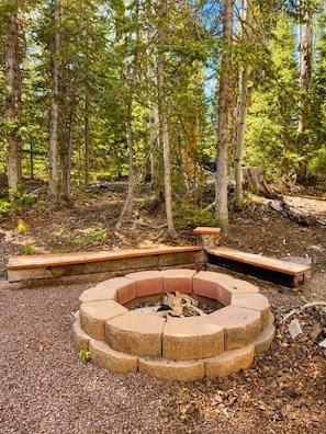 A circular fire pit surrounded by stone seating, set in a forested area with trees and a clear sky. - Mountain View Hideaway (Brian Head)