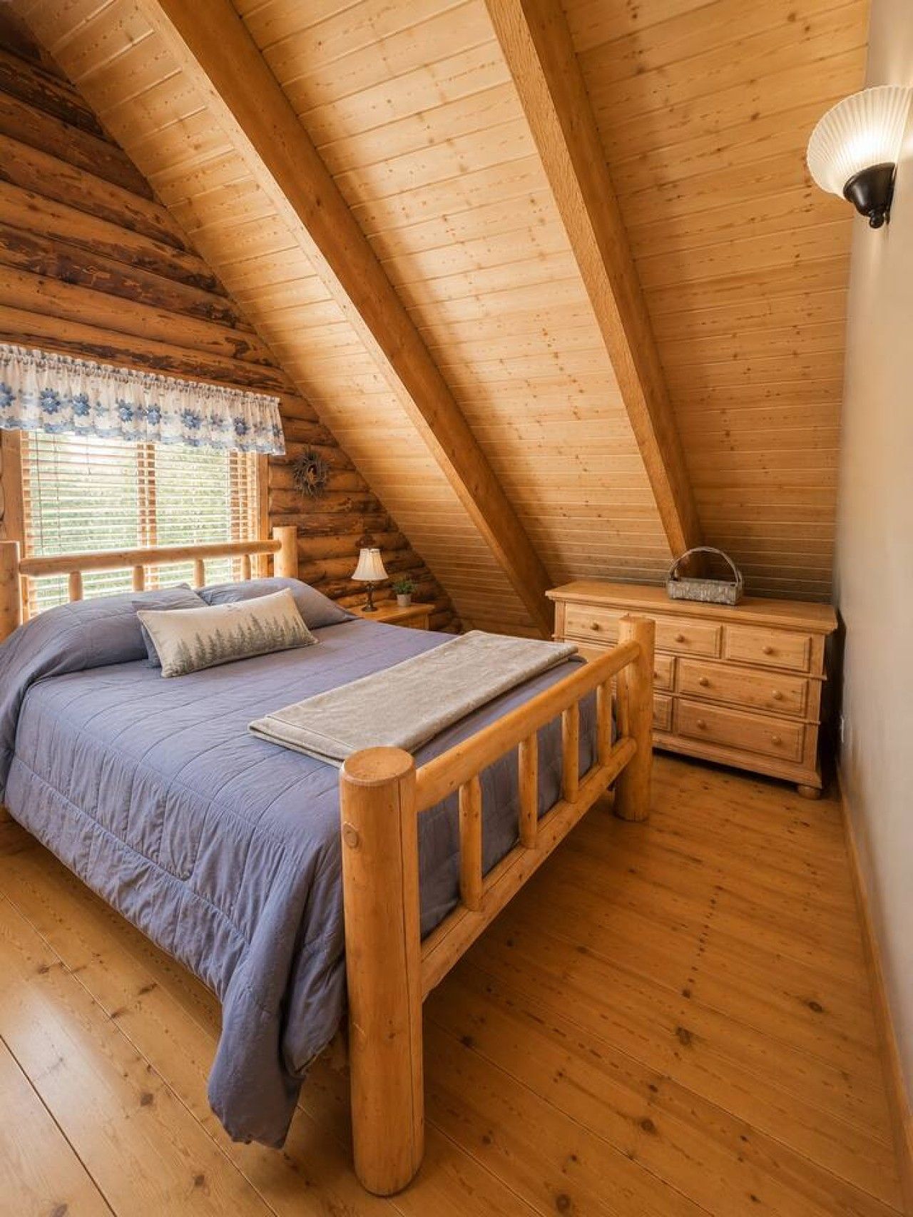A cozy attic bedroom with a wooden bed, a nightstand, a dresser, and a window with blinds.