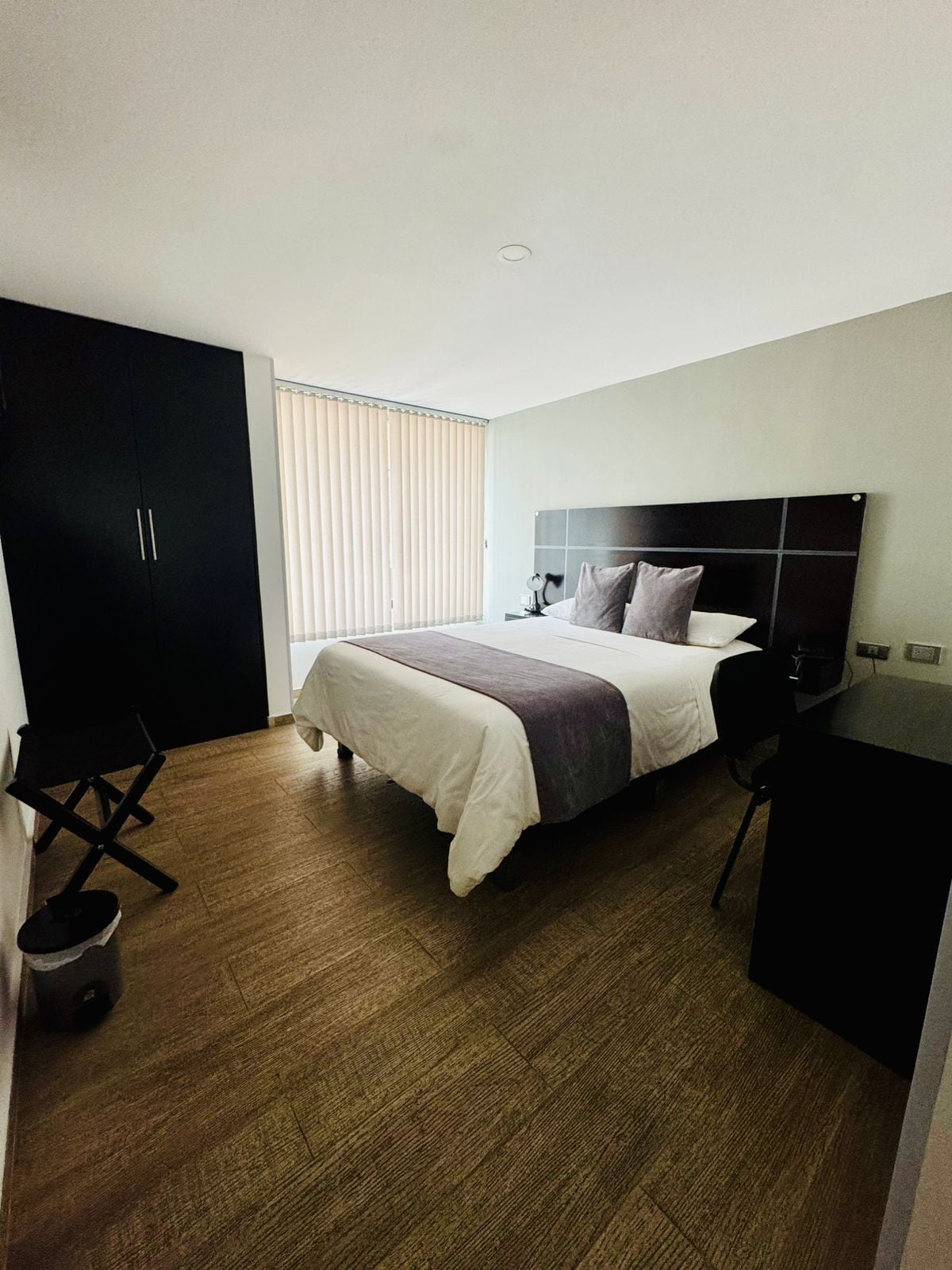 A hotel room with a bed, a desk, a wardrobe, and a window with blinds.
