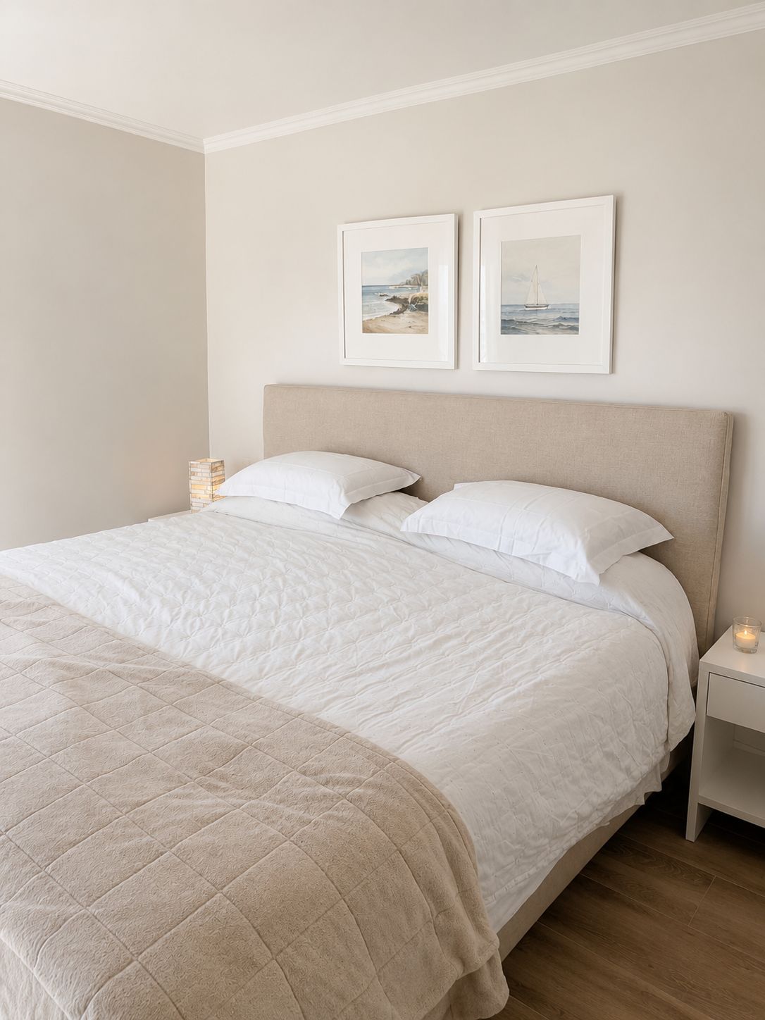 A neatly made bed with a beige headboard, white bedding, and a quilted blanket. There are two framed pictures on the wall above the bed.