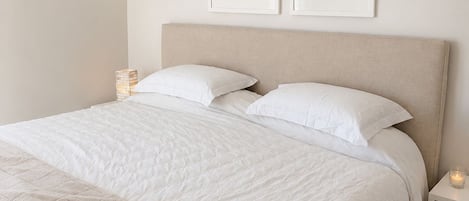 A neatly made bed with a beige headboard, white bedding, and a quilted blanket. There are two framed pictures on the wall above the bed.