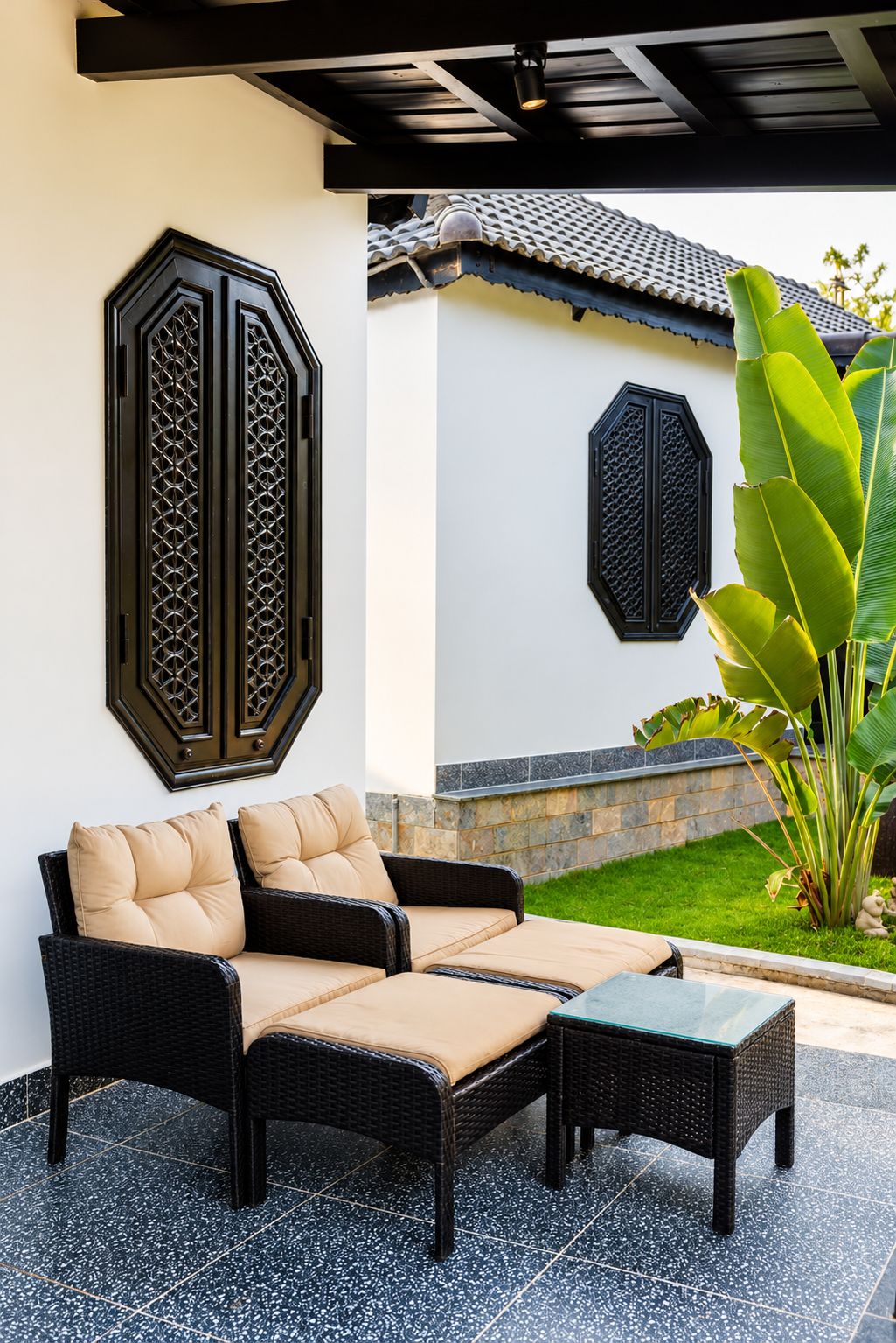 A patio area with a wicker sofa and matching ottoman, a decorative wall panel, and a large plant.