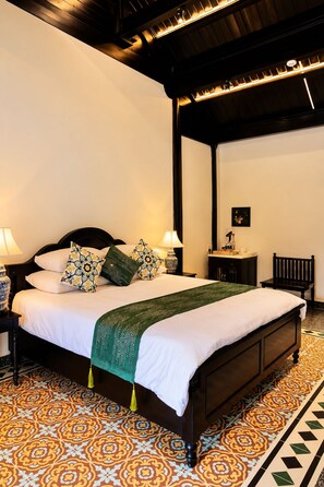 Cama bem arrumada com manta verde, mesa de cabeceira com abajur e cadeira em um quarto com ladrilhos estampados.