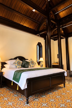Quarto de hotel com uma cama grande, vigas de madeira no teto, piso estampado e mesa de cabeceira com abajur.
