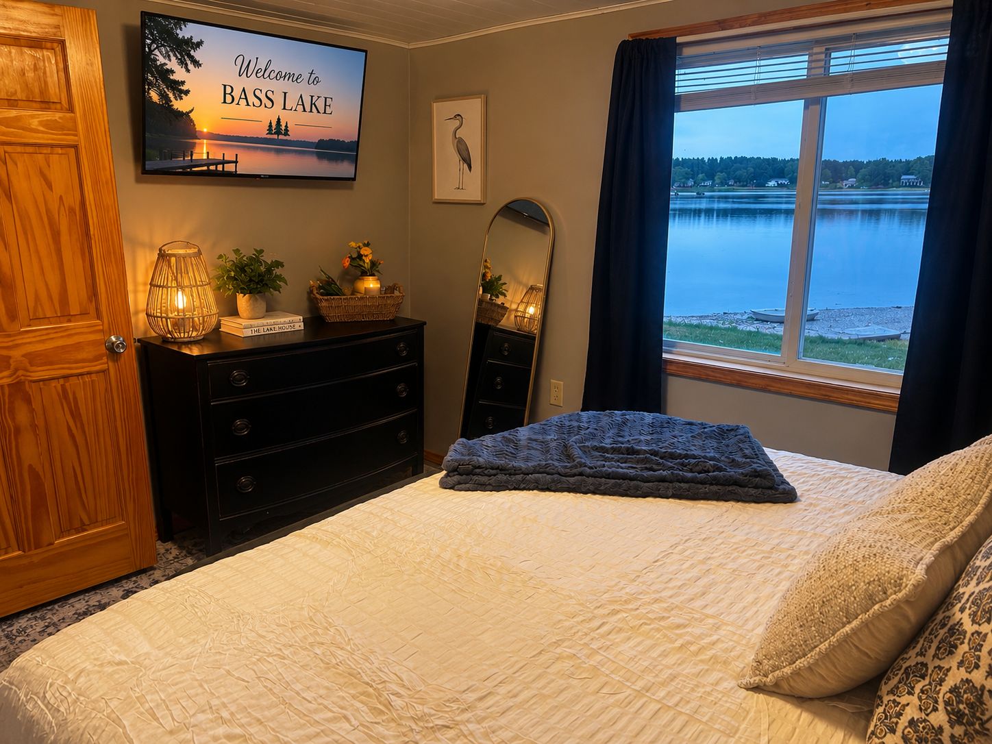 Une chambre à coucher comprenant un lit, une commode, un téléviseur affichant un message de bienvenue, une fenêtre donnant sur un lac et un tableau encadré représentant un oiseau.