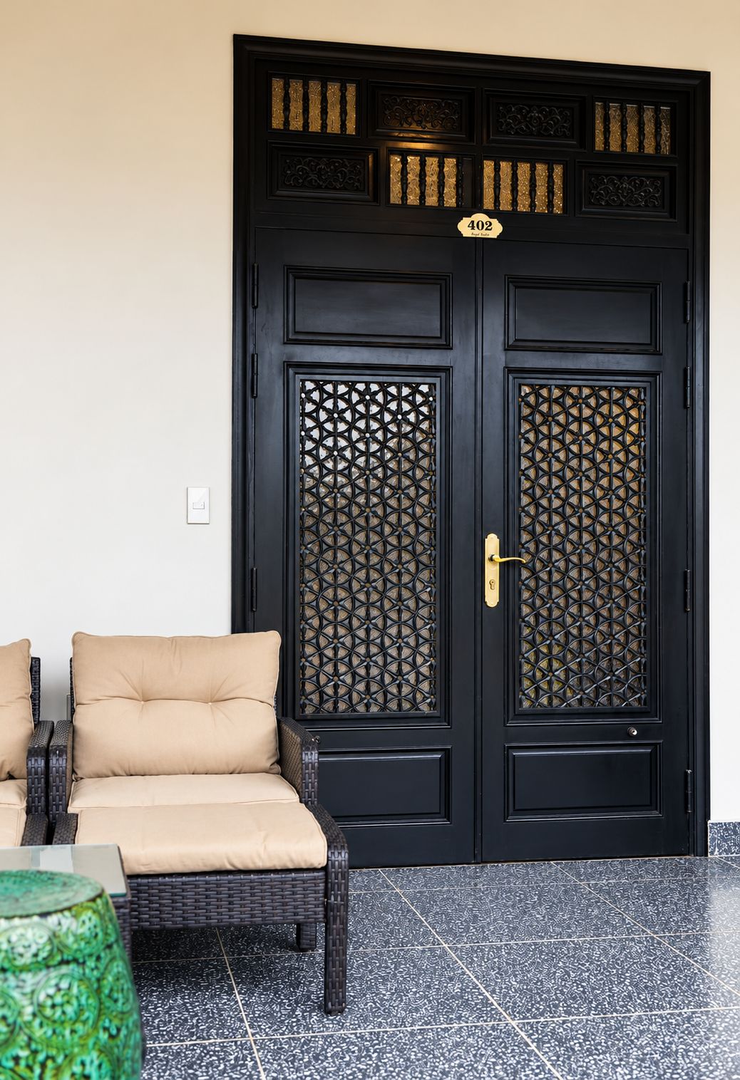 A black double door with intricate patterns and a gold number plate, a beige sofa, and a green vase.