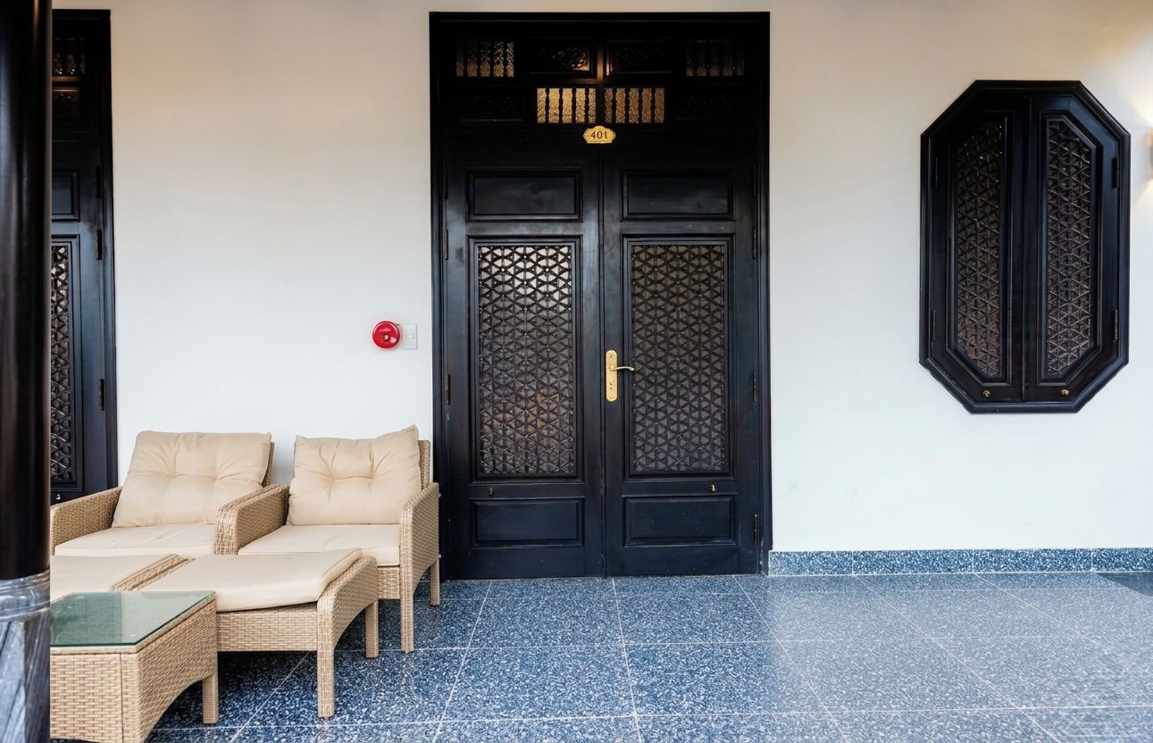 A black double door with a number plate, a red fire alarm, and a wicker sofa with cushions.