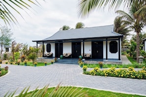 Um edifício de dois andares com telhado de telhas, varanda coberta com assentos e jardim ajardinado com palmeiras e flores.
