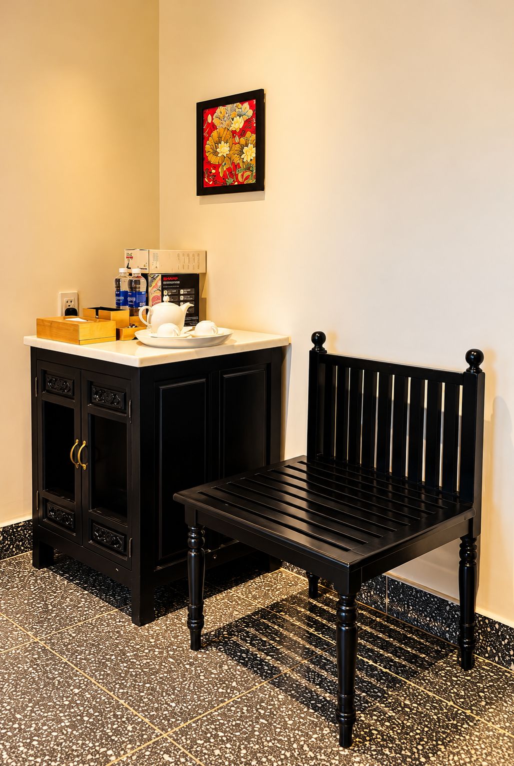 A room with a black chair, a small table with a white cup, and a cabinet with a framed picture above it.