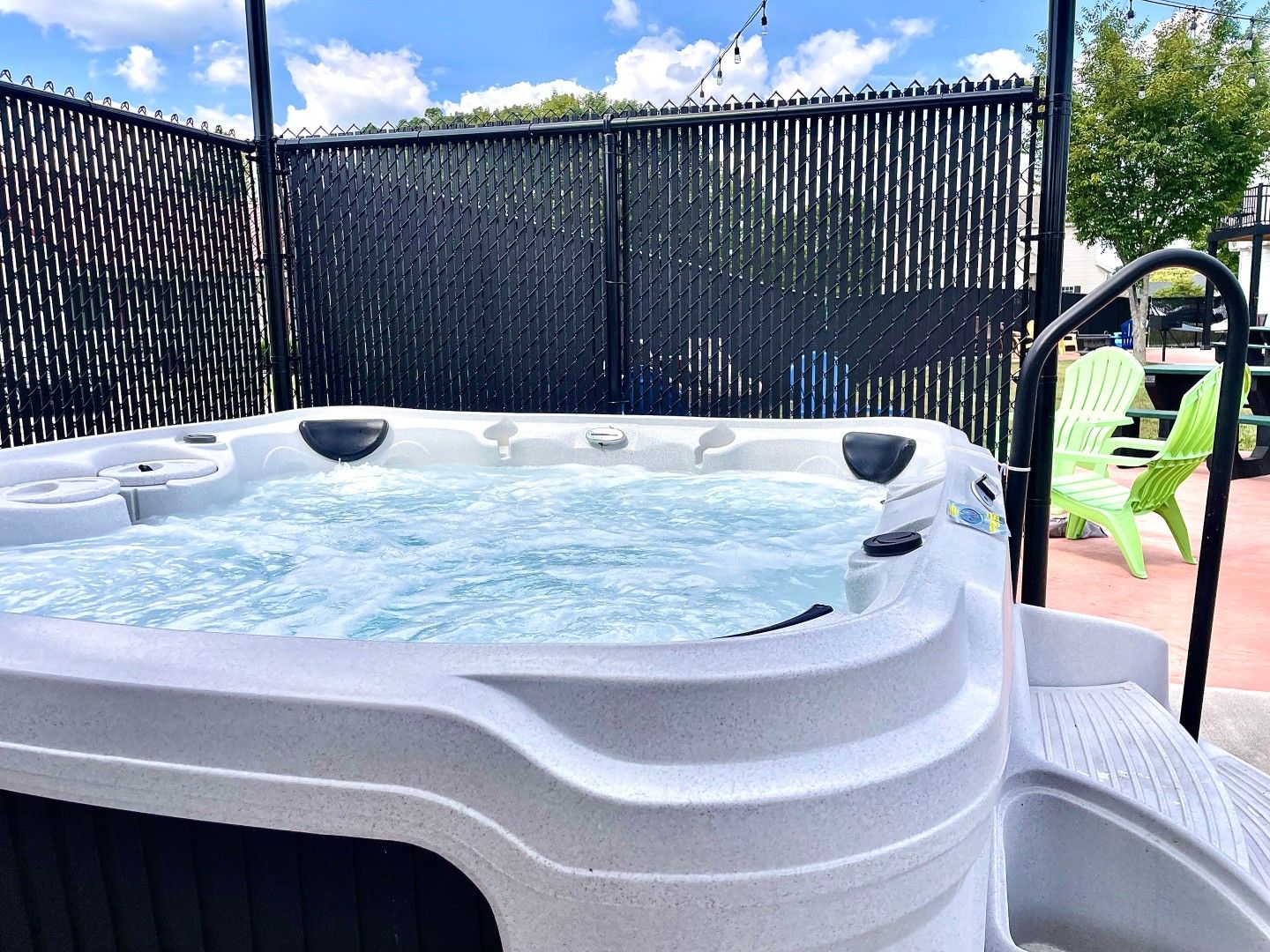 A hot tub with a black fence enclosure, a green chair, and a patio area.