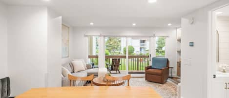 A modern living room with a wooden dining table, a sofa, and a chair. There is a balcony with a view of the outside.