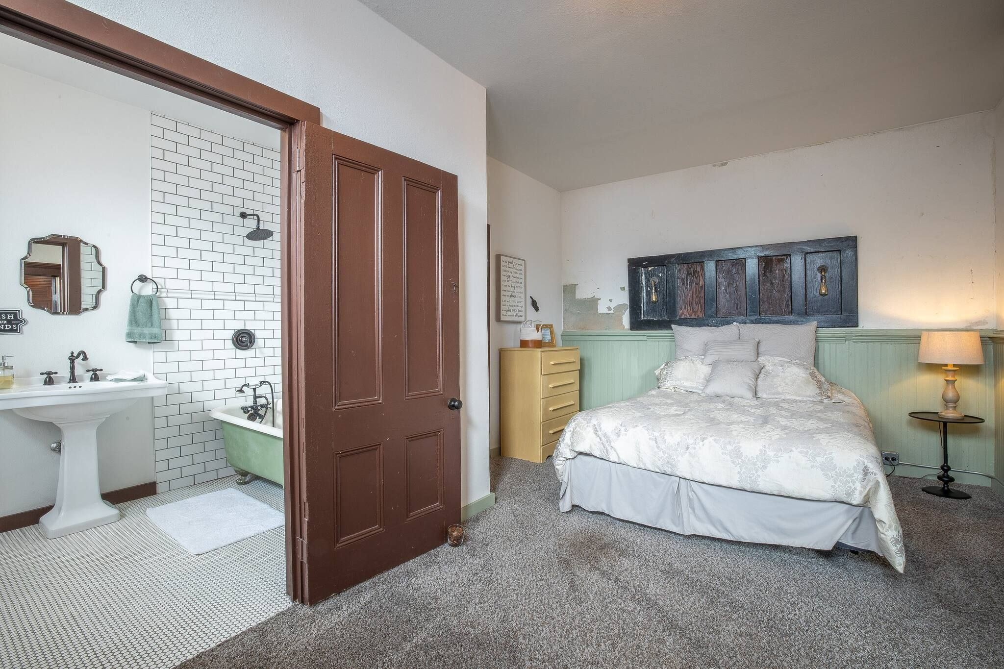 Ein Schlafzimmer mit Bett, Kommode, Nachttisch, Lampe, Spiegel, Tür und einem Badezimmer mit Waschbecken und Badewanne.