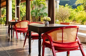 Een overdekt terras met rode stoelen en tafels, omgeven door groen.