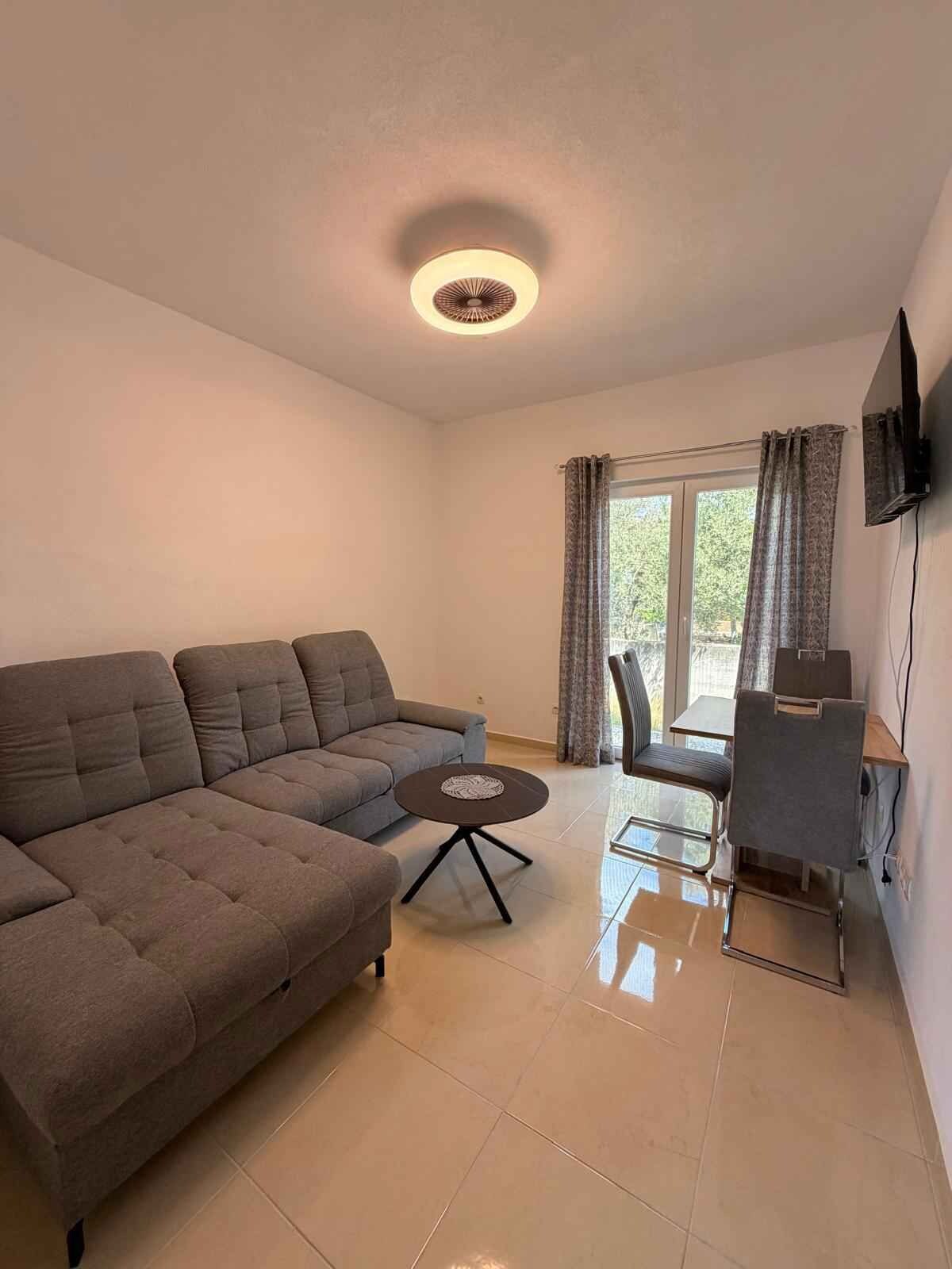 A living room with a sectional sofa, a small round table, a dining table with chairs, a television mounted on the wall, and a window with curtains.