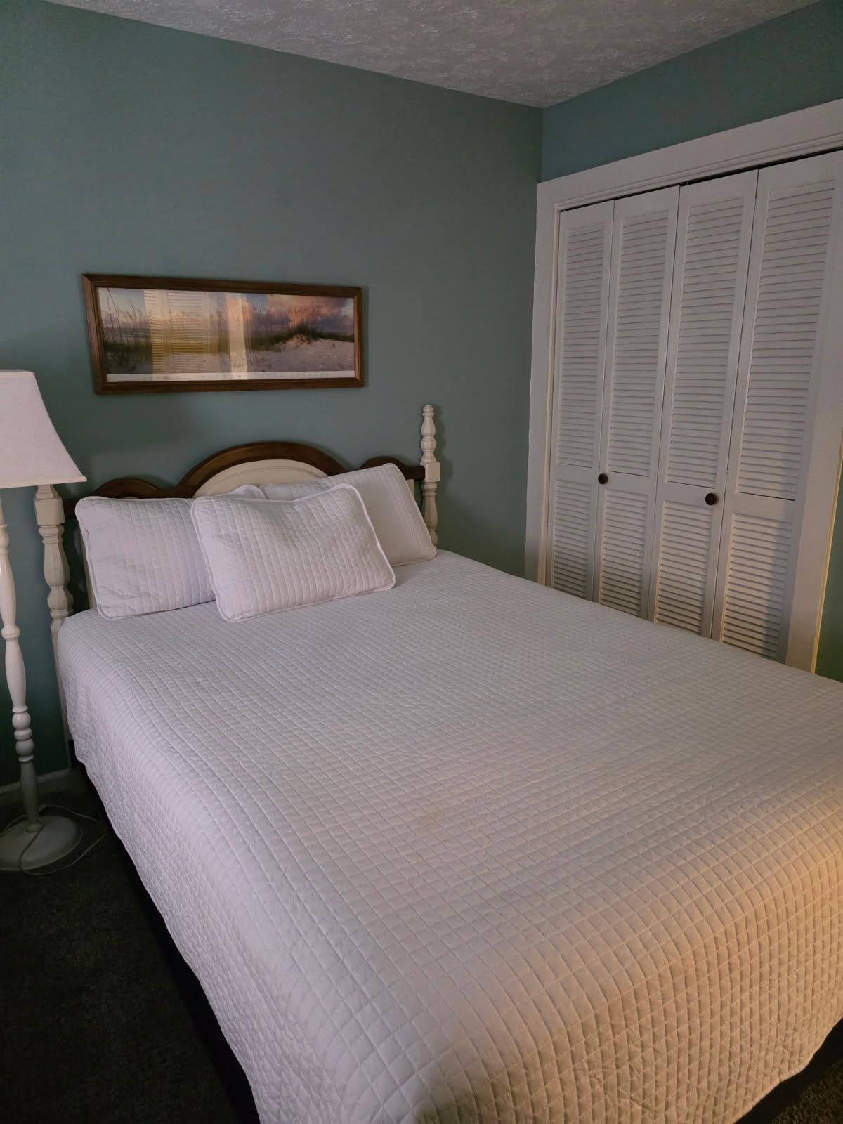 A bedroom with a bed, a nightstand, a lamp, a painting, and a set of louvered doors.
