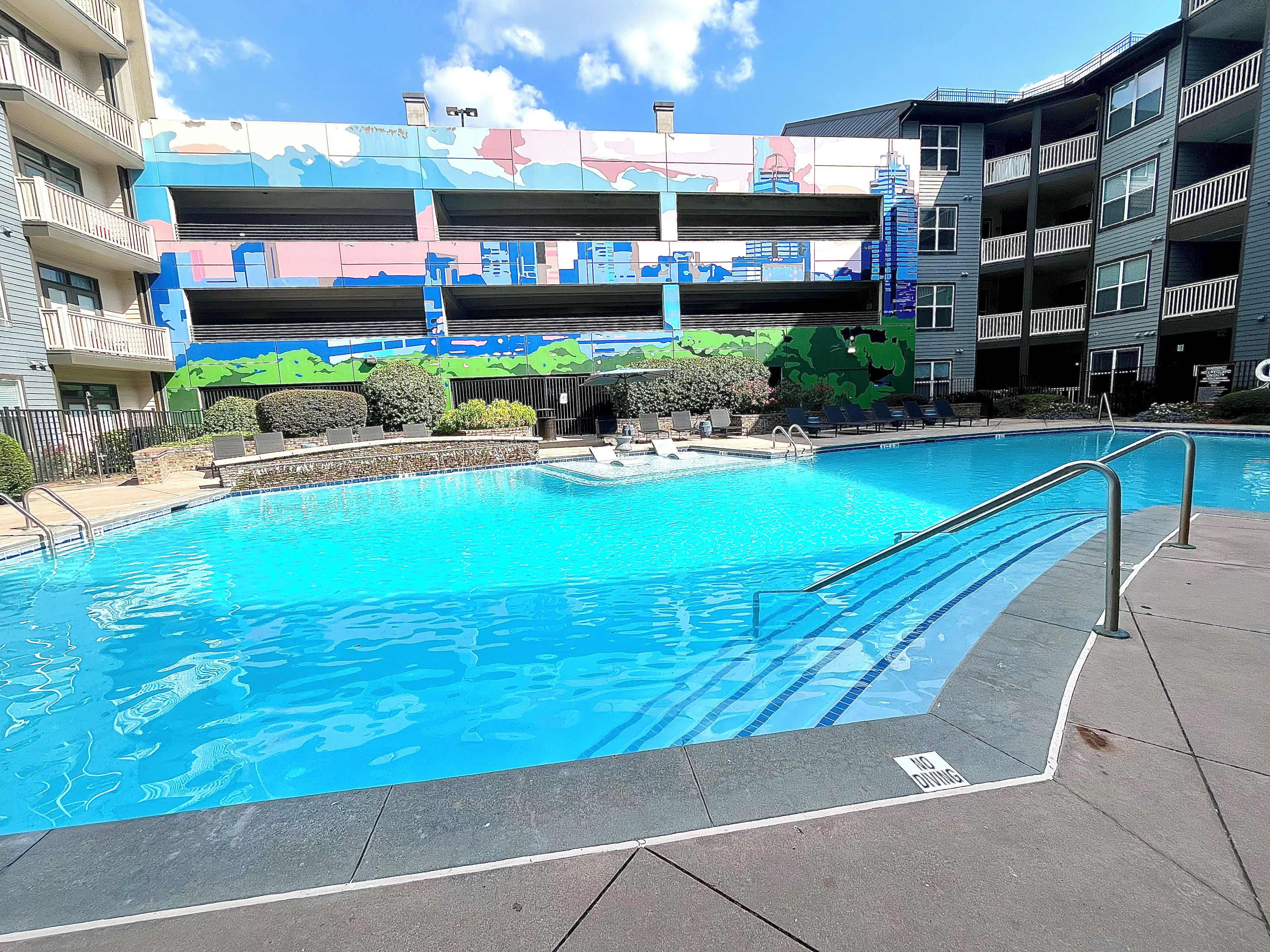 A swimming pool with a no diving sign, surrounded by a landscaped area with lounge chairs and a multi-story building with colorful murals in the background.