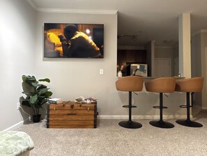 Ein Wohnzimmer mit an der Wand montiertem Fernseher, einer Holzkommode, einem Blumentopf und drei Barhocker.