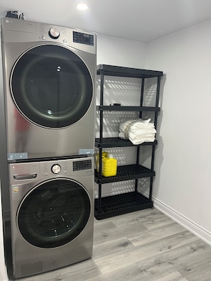 A laundry area with a stacked washer and dryer, a shelf with folded clothes, and a bottle of detergent. - Welcome to Skies Abode. (Brant)