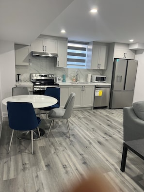 A modern kitchen with white cabinets, stainless steel appliances, a dining table with blue chairs, and a grey sofa. - Welcome to Skies Abode. (Brant)