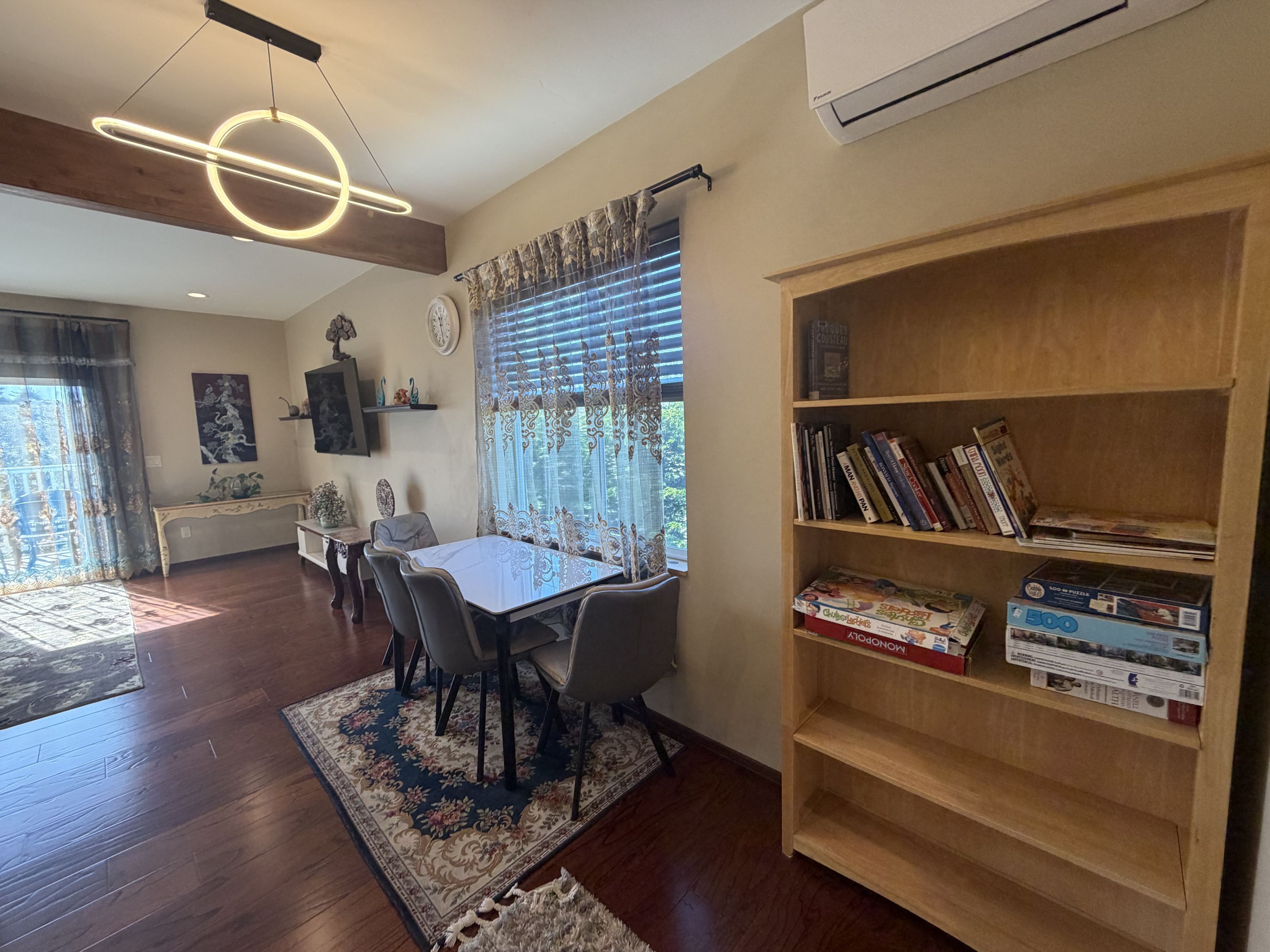 A dining area with a table and chairs, a bookshelf with books and board games, a clock on the wall, and a window with curtains.