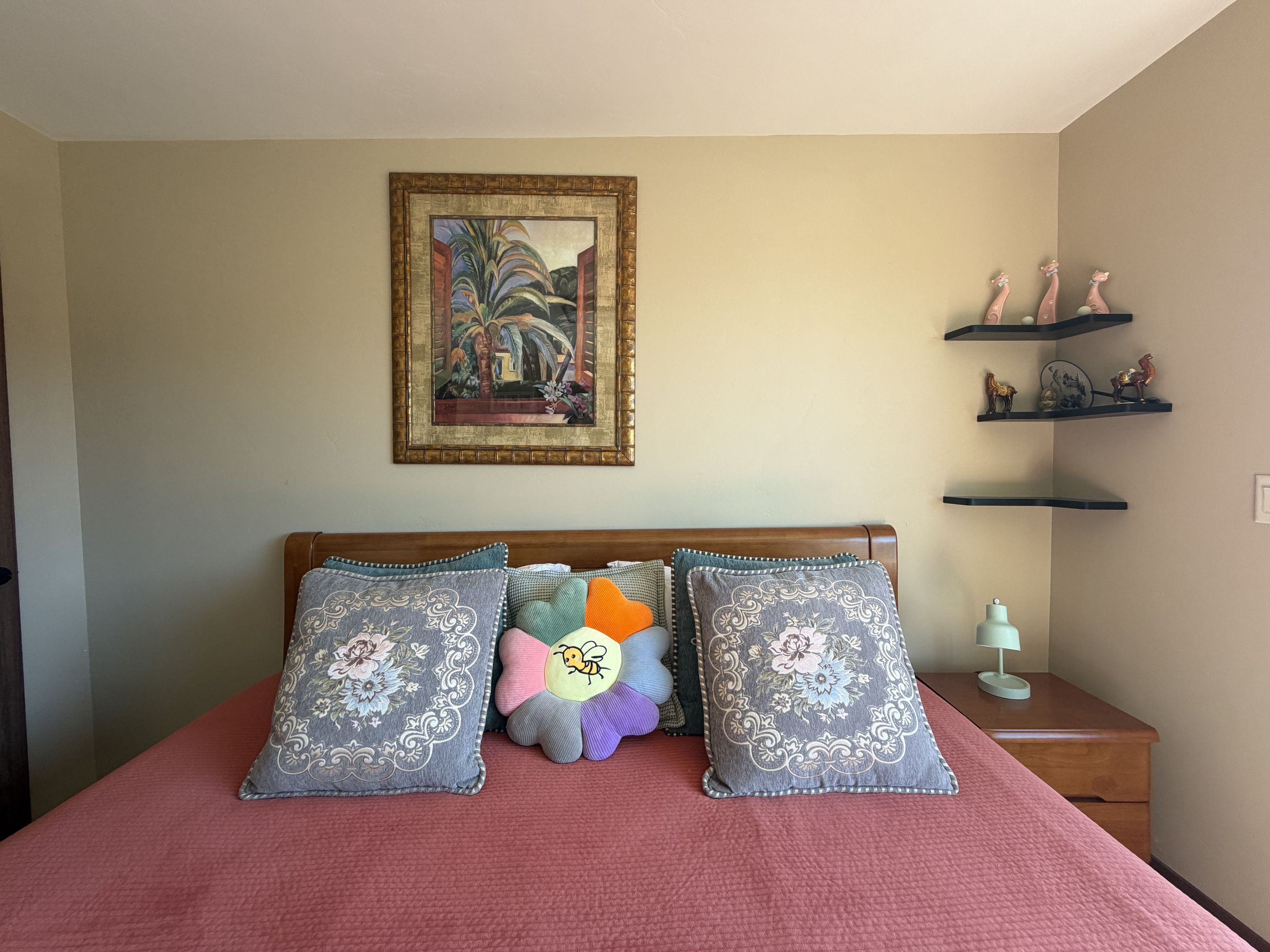 A bedroom with a bed, pillows, a nightstand, a lamp, and a wall-mounted shelf with decorative items.