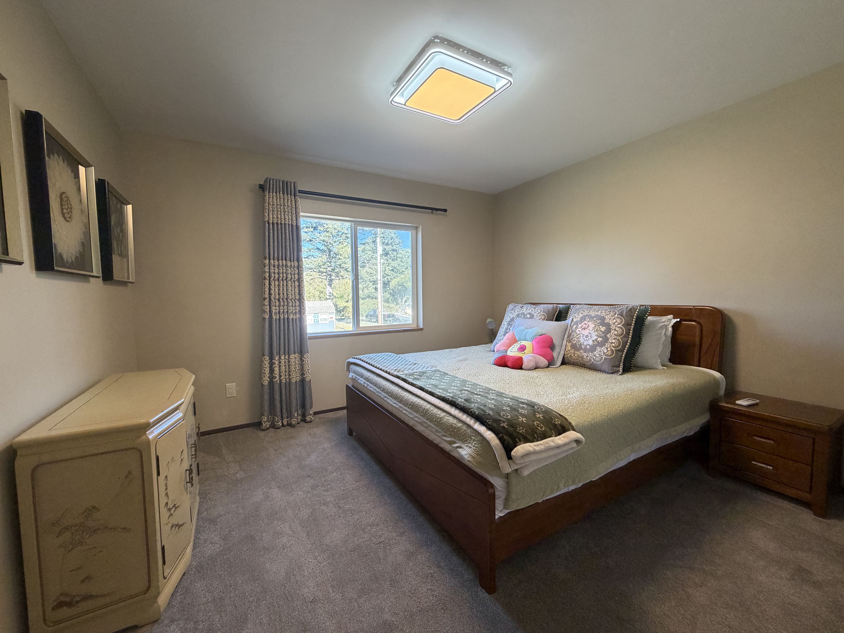 A bedroom with a bed, a nightstand, a dresser, a window with curtains, and a ceiling light.