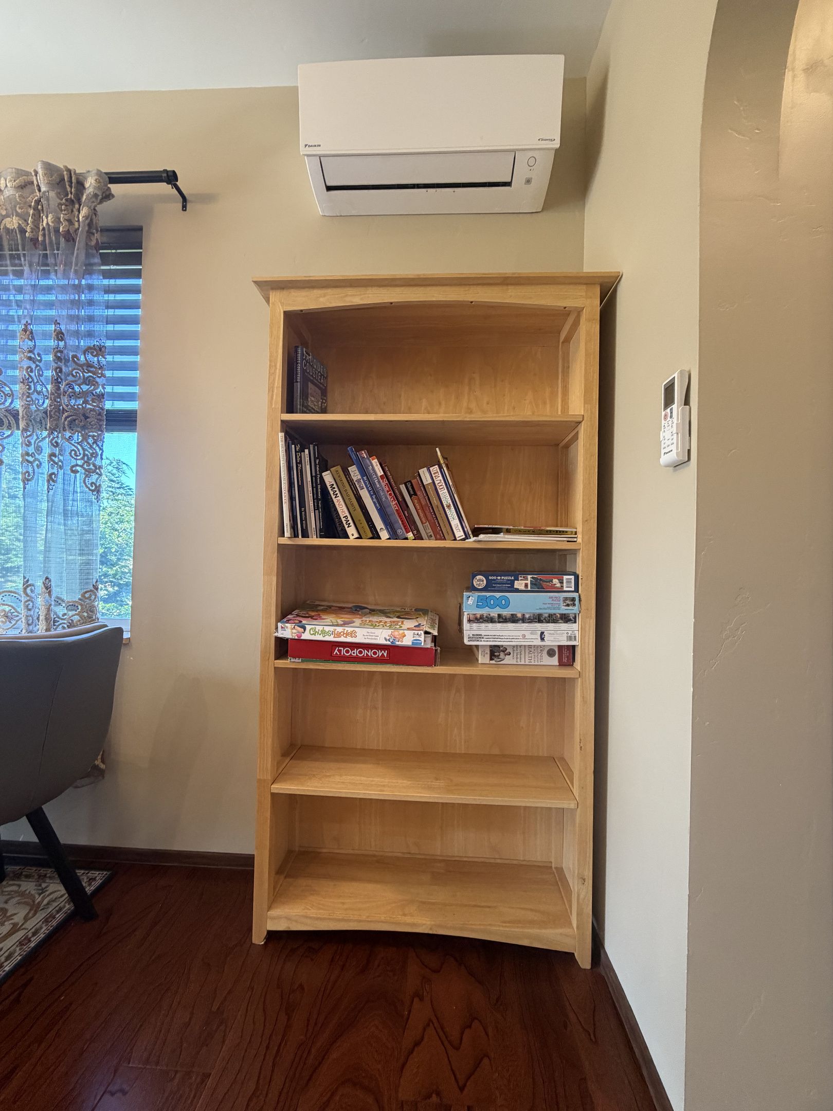 A room with a wooden bookshelf, a window with curtains, and an air conditioning unit mounted on the wall.