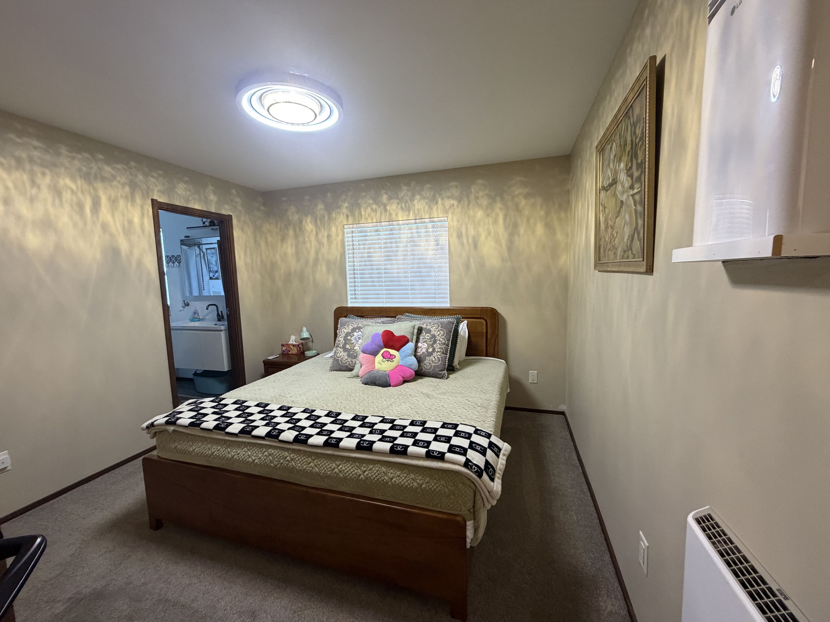 A bedroom with a bed, a nightstand, a window with blinds, a radiator, and a door leading to a bathroom.