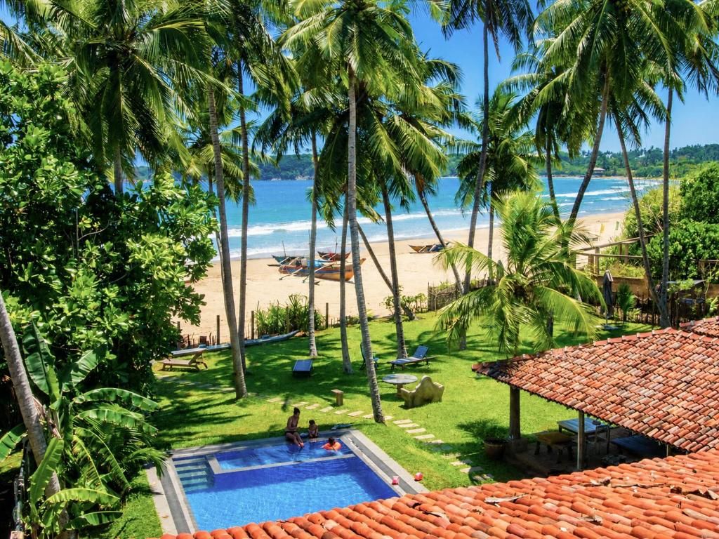 Zona de piscina con vista a la playa y al océano, rodeada de palmeras y una estructura de techo rojo.
