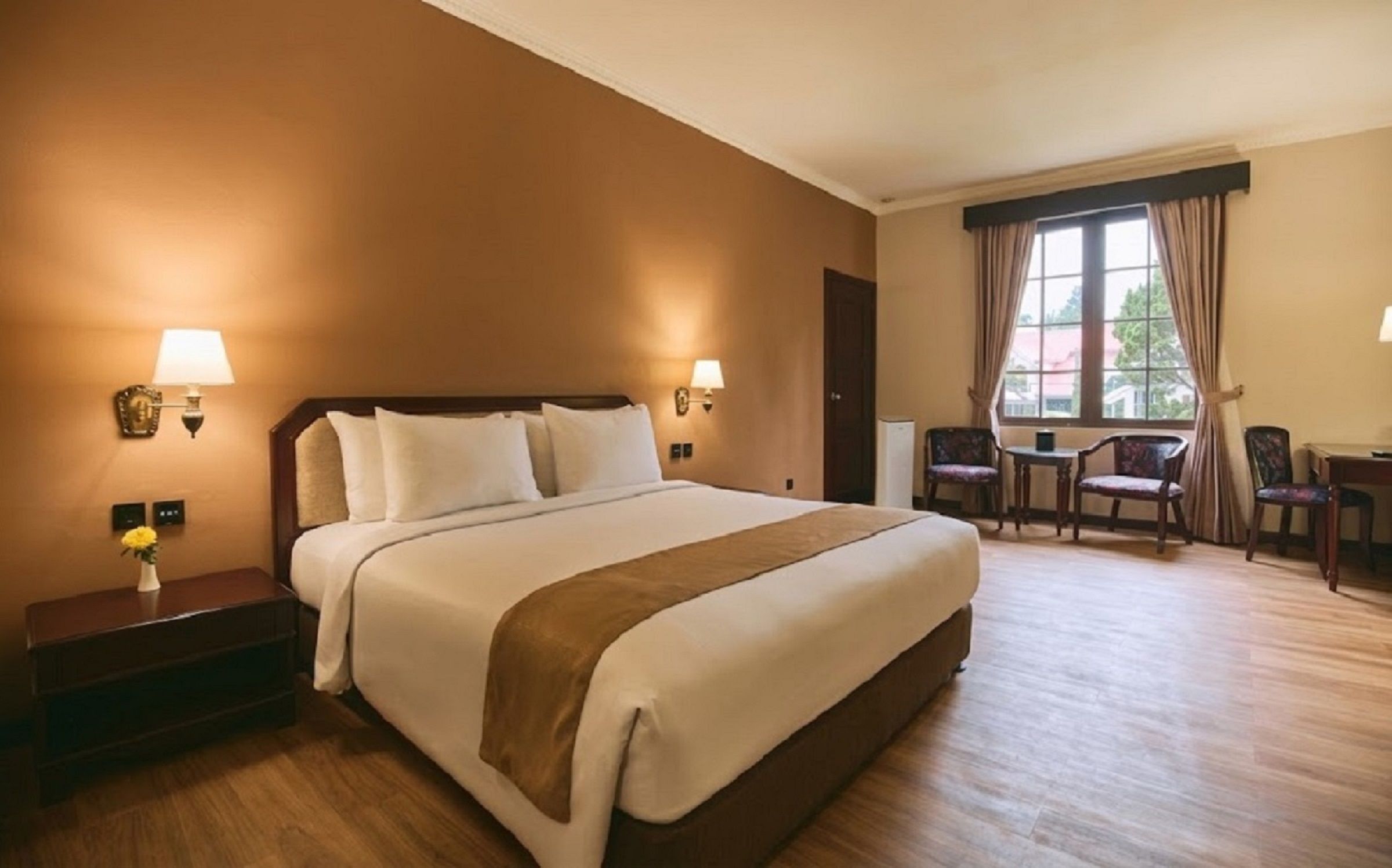 A hotel room with a large bed, bedside lamps, a desk with a chair, and a window with curtains.