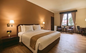 A hotel room with a large bed, bedside lamps, a desk with a chair, and a window with curtains.