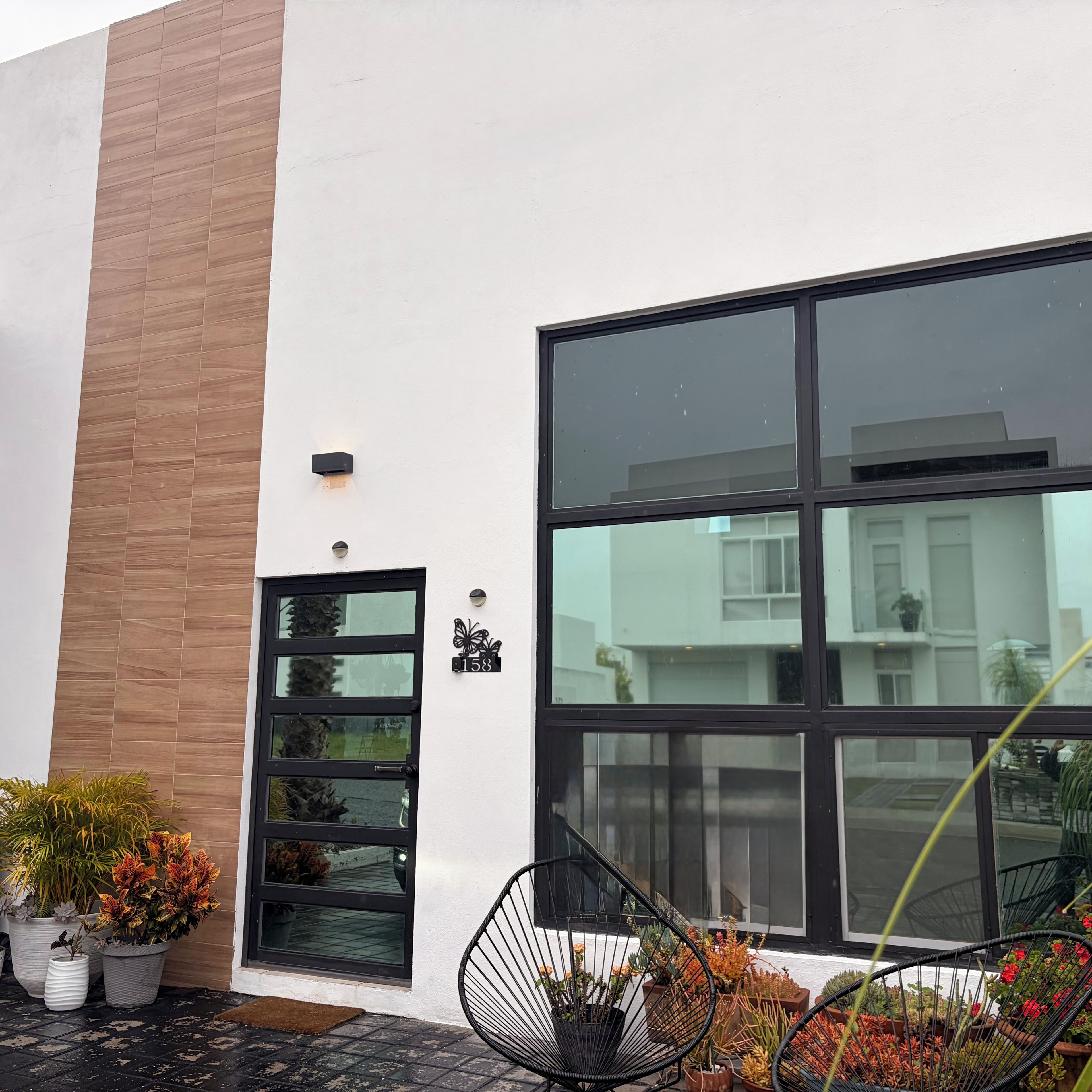 Un bâtiment moderne doté d’une grande fenêtre et d’une porte, avec des plantes en pot et une chaise à l’extérieur.