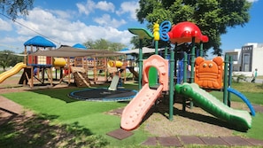 Un parque infantil con diversas estructuras de juego, incluyendo toboganes, una zona de escalada y un pequeño edificio con techo azul.