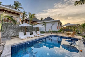 A swimming pool with lounge chairs and umbrellas, surrounded by a stone wall and palm trees.