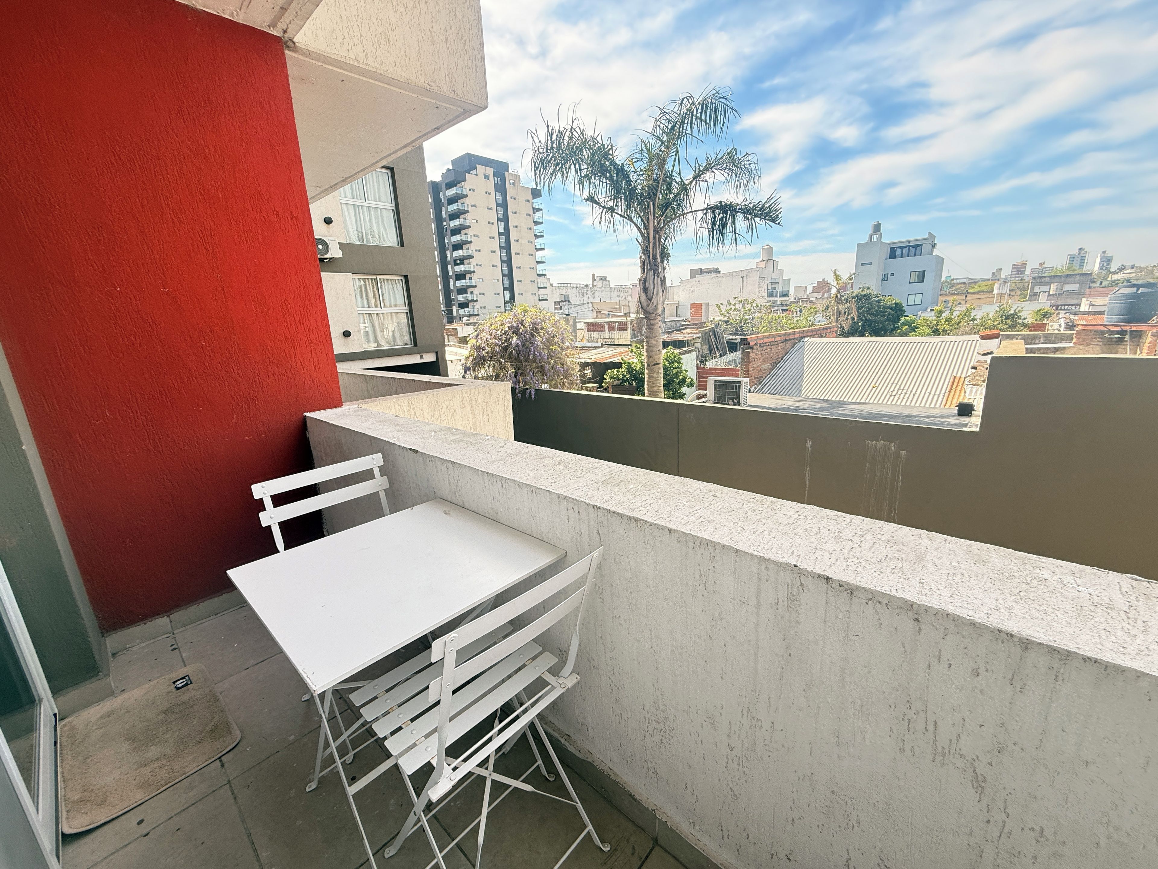 Varanda com mesa e cadeira brancas, parede vermelha e vista para um horizonte urbano com edifícios e palmeiras.