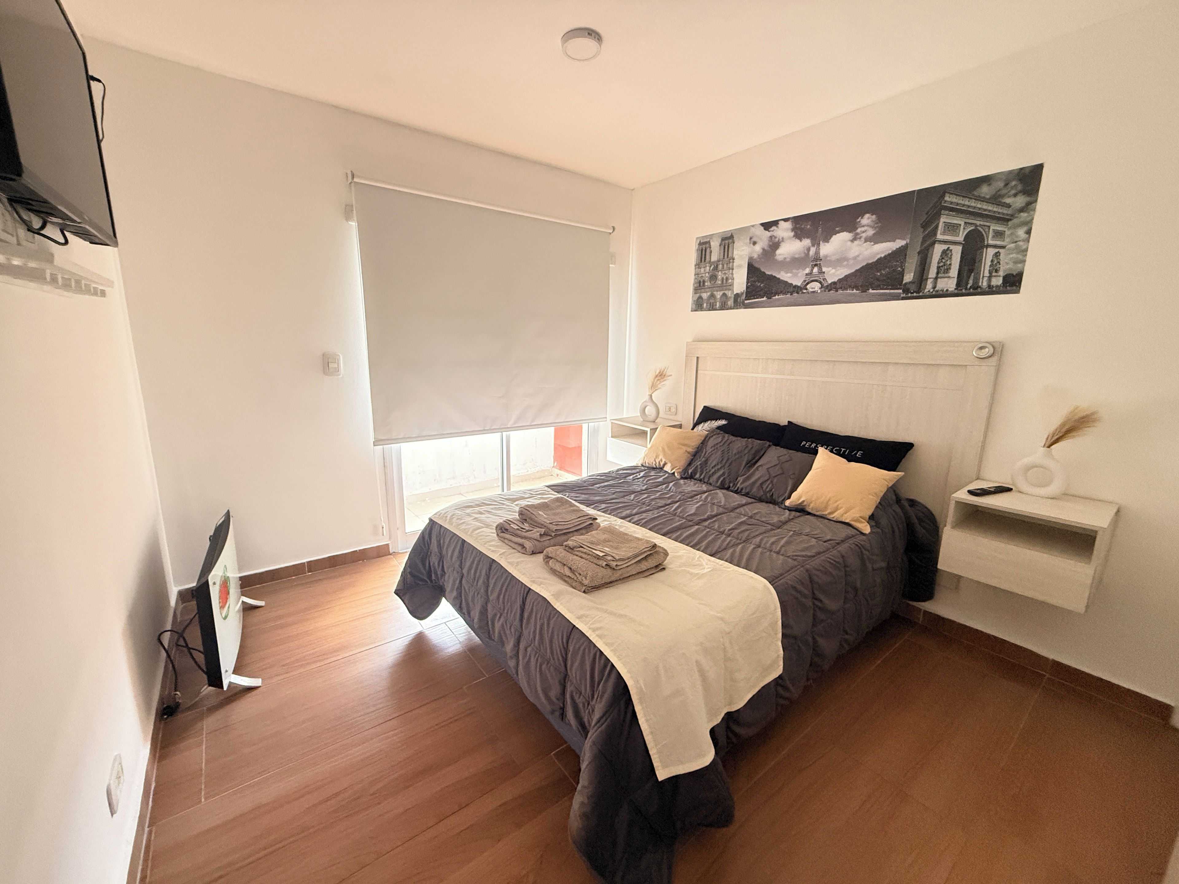 Quarto com cama, janela com persiana, quadro na parede e mesa de cabeceira com controle remoto e um vaso.